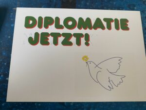 Postkarte der UGL mit einer stilisierten Friedenstaube, die an diejenige Pablo Picassos erinnert, und dem Text "Diplomatie jetzt!"